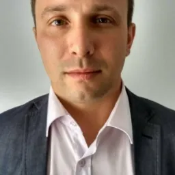 Portret mężczyzny w białej koszuli i szarej marynarce na jasnym tle, z widocznymi detalami twarzy i delikatnym uśmiechem.