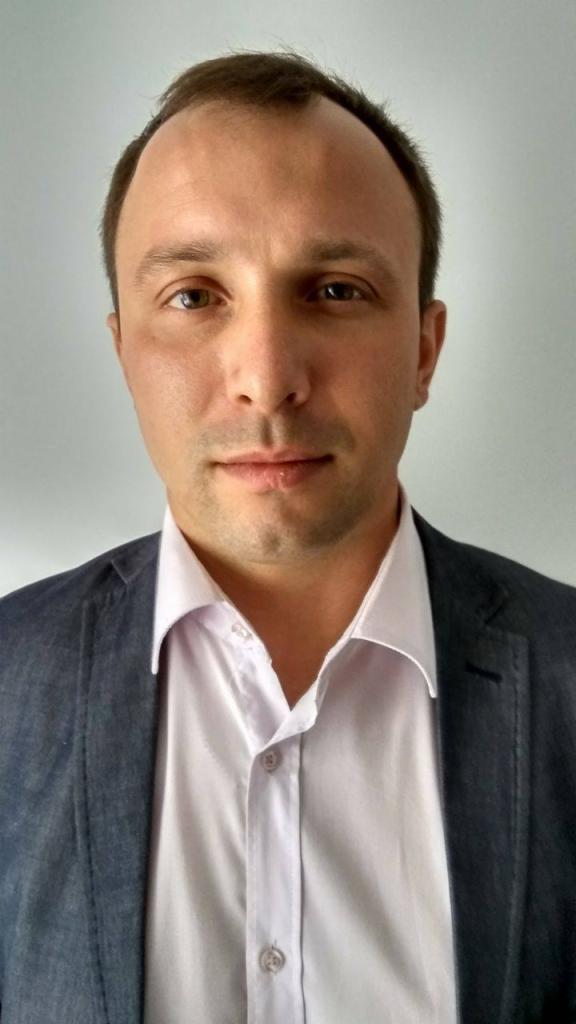 Portret mężczyzny w białej koszuli i szarej marynarce na jasnym tle, z widocznymi detalami twarzy i delikatnym uśmiechem.