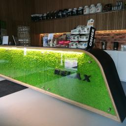 Nature's Design - Produkty Natury Claus Sternahl - Recepcja z frontem dekorowanym mchem, podświetlona od góry, z napisem 'RELAX' w czarnych literach. Na półkach za ladą widoczne suplementy diety i napoje.