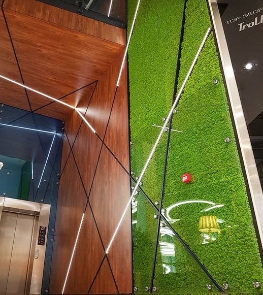 Nowoczesna ściana z mchu w biurze, z drewnianymi panelami i liniowym oświetleniem LED, widoczny fragment windy i logo firmy w tle.