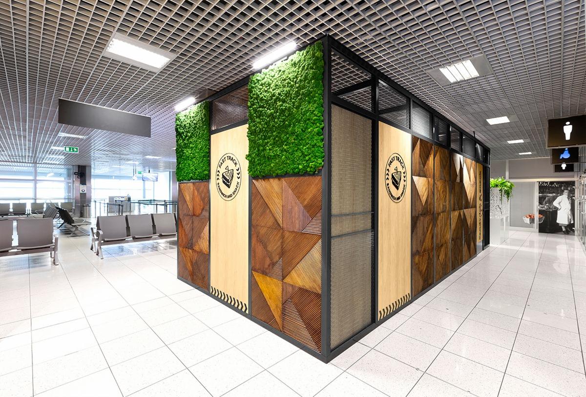 Nowoczesna aranżacja przestrzeni komercyjnej z panelami drewnianymi o geometrycznym wzorze, pionowymi panelami z mchu oraz logo 'Fast Track Sandwich and Snack', w jasnym wnętrzu z białą podłogą...
