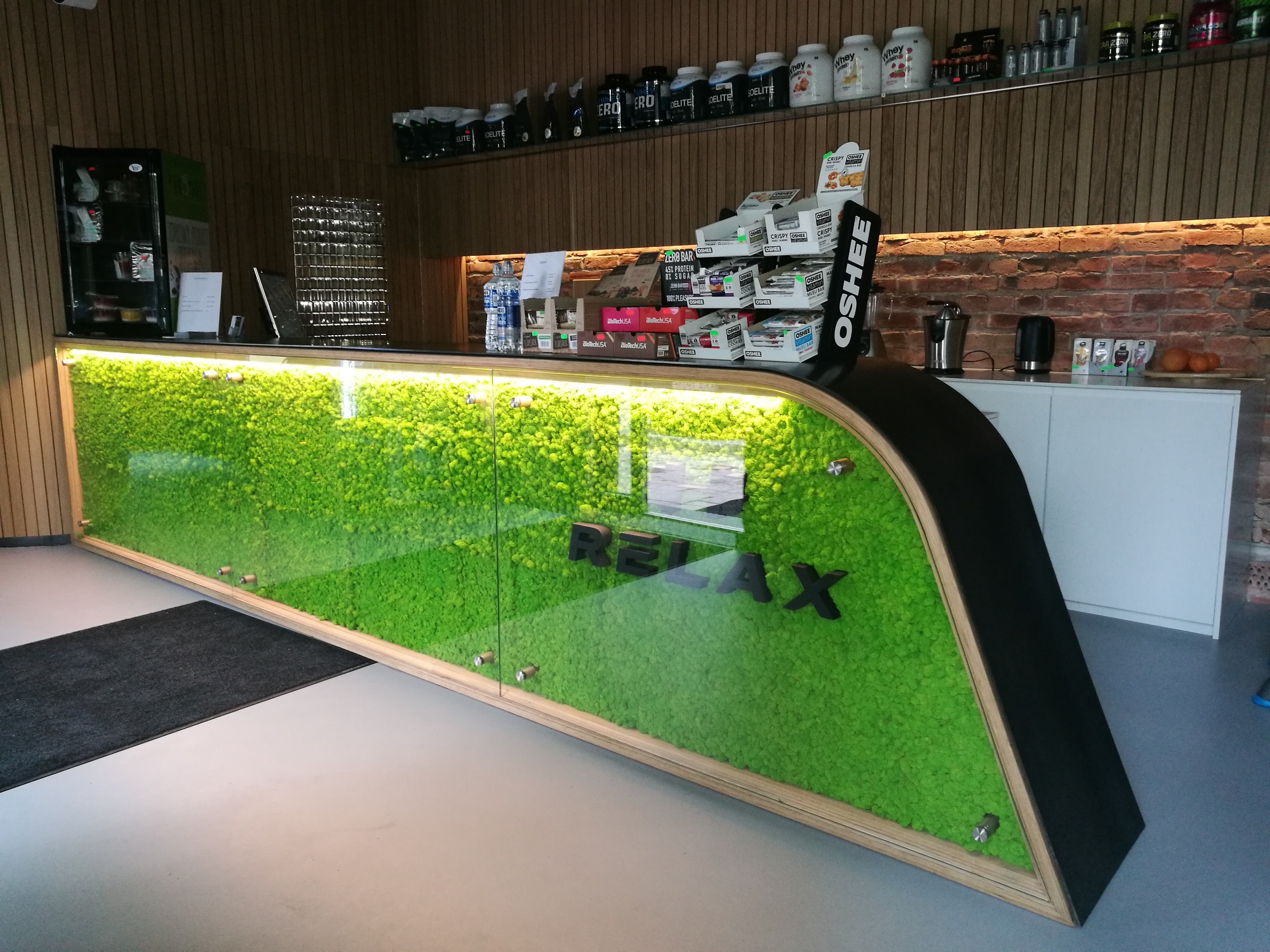 Recepcja z frontem dekorowanym mchem, podświetlona od góry, z napisem 'RELAX' w czarnych literach. Na półkach za ladą widoczne suplementy diety i napoje.