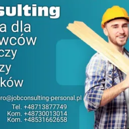 Mężczyzna w kasku budowlanym i koszuli w kratę, uśmiechnięty, trzymający na ramieniu deski, na tle reklamy z ofertami pracy dla spawaczy, murarzy i hydraulików oraz informacją o stawkach godzinowych.