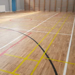 Sala gimnastyczna z jasną, drewnianą podłogą parkietową, pomalowaną w kolorowe linie boiska, z drabinkami gimnastycznymi przy ścianie.