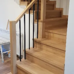 Jasne drewniane schody z czarnymi metalowymi balustradami, widoczne niedokończone wykończenie ściany i podłogi, sugerujące etap montażu.