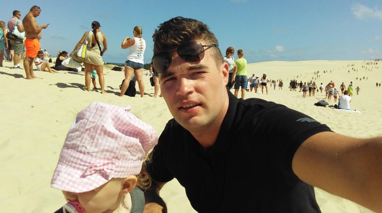 Selfie mężczyzny z dzieckiem w różowej czapeczce na tle piaszczystej wydmy z tłumem ludzi w słoneczny dzień.