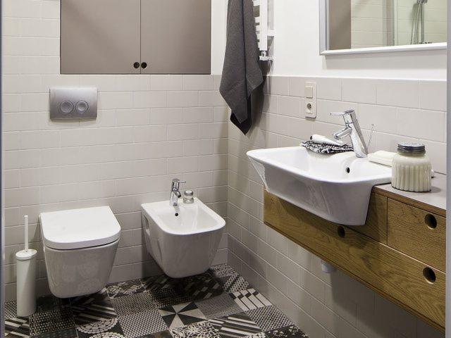 Nowoczesna łazienka z białymi płytkami, geometryczną posadzką, wiszącą toaletą i bidetem, umywalką na drewnianej szafce oraz szarym ręcznikiem.