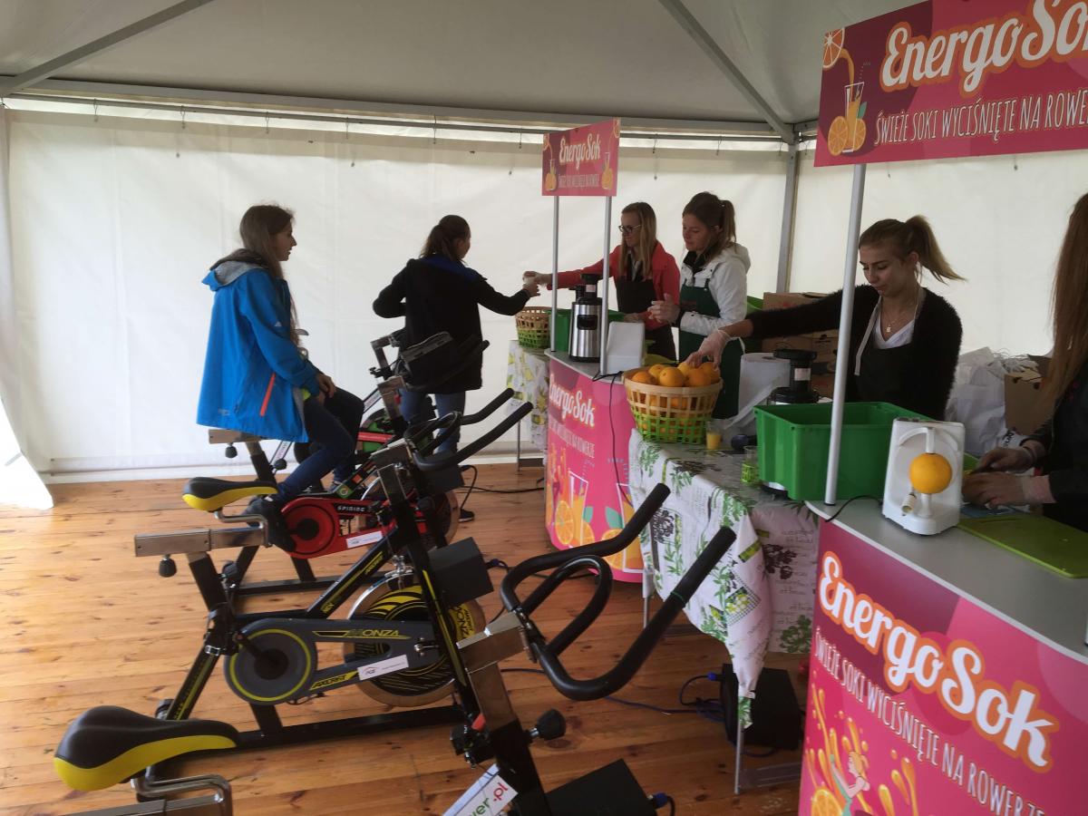 Stoisko z wyciskanymi sokami 'Energo Sok' z rowerami stacjonarnymi do napędzania wyciskarki. Kobieta w niebieskiej kurtce siedzi na rowerze, a trzy osoby obsługują stoisko z pomarańczami...