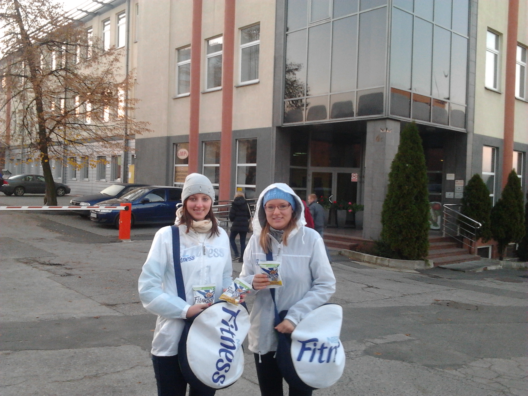 Dwie uśmiechnięte kobiety w kurtkach przeciwdeszczowych i czapkach prezentują paczki przekąski Fitness, stojąc na zewnątrz budynku z przeszkloną fasadą i zielonymi krzewami.
