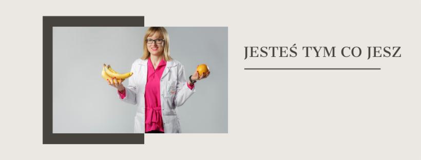 Uśmiechnięta kobieta w okularach i białym fartuchu lekarskim trzyma w dłoniach owoce: banany i pomarańczę, obok napis 'Jesteś tym co jesz'.