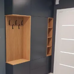 Ciemnoszara szafa na wymiar z drewnianymi elementami: wieszaki na ubrania i półki, przylegająca do białych drzwi.