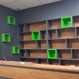 Szara ściana z drewnianymi panelami i półkami, w tym kilka jaskrawo zielonych, w trakcie montażu, na blacie poziomica laserowa.