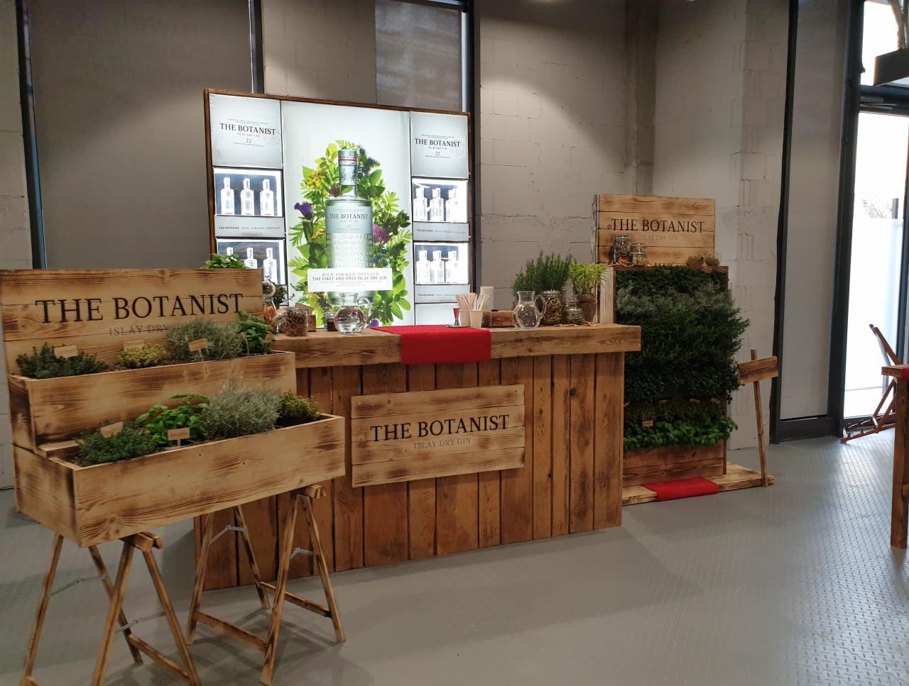 Drewniany bar z ziołami i logiem 'The Botanist' w industrialnym wnętrzu. Zioła w skrzynkach, butelka na podświetlonym tle. Elementy dekoracyjne z drewna.