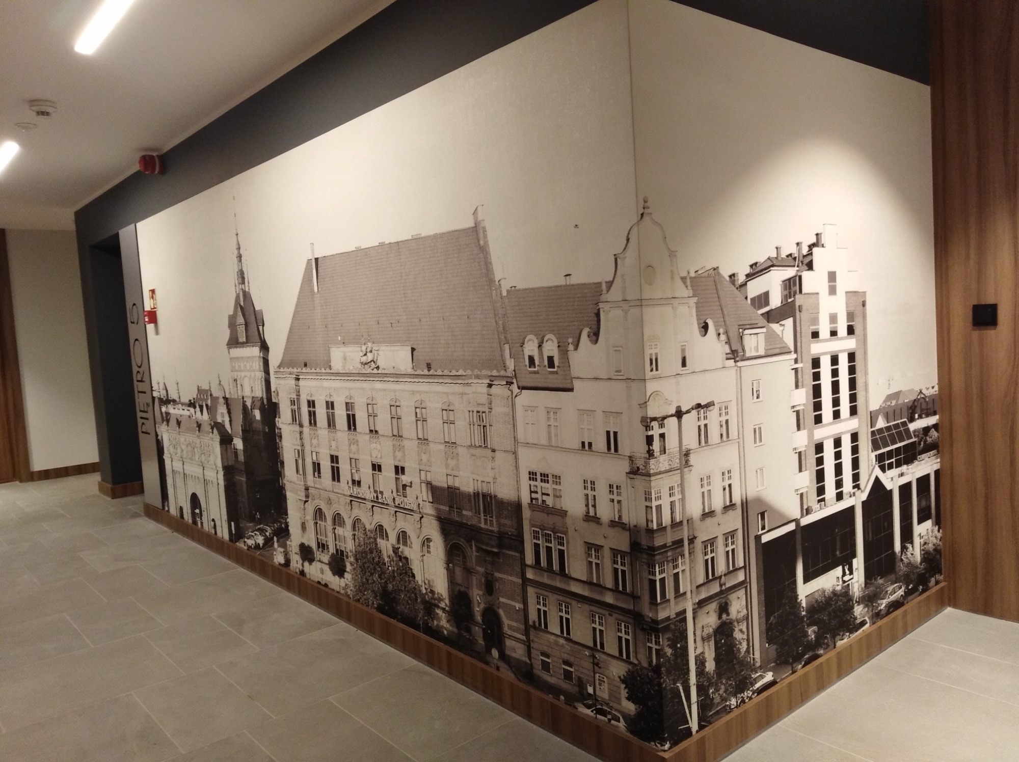 Tapeta z motywem czarno-białej panoramy miejskiej z historyczną architekturą, na ścianie korytarza z szarą podłogą i drewnianymi elementami wykończeniowymi.