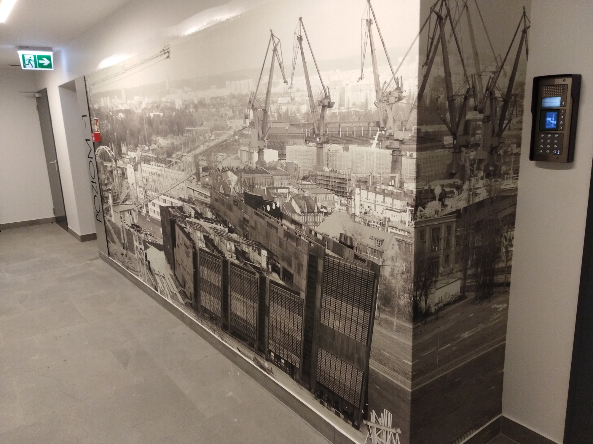 Fototapeta w korytarzu w Gdyni przedstawia czarno-biały widok na stocznię z dźwigami, tworząc industrialny klimat. Na ścianie widoczny domofon, a obok fototapety znak ewakuacyjny.