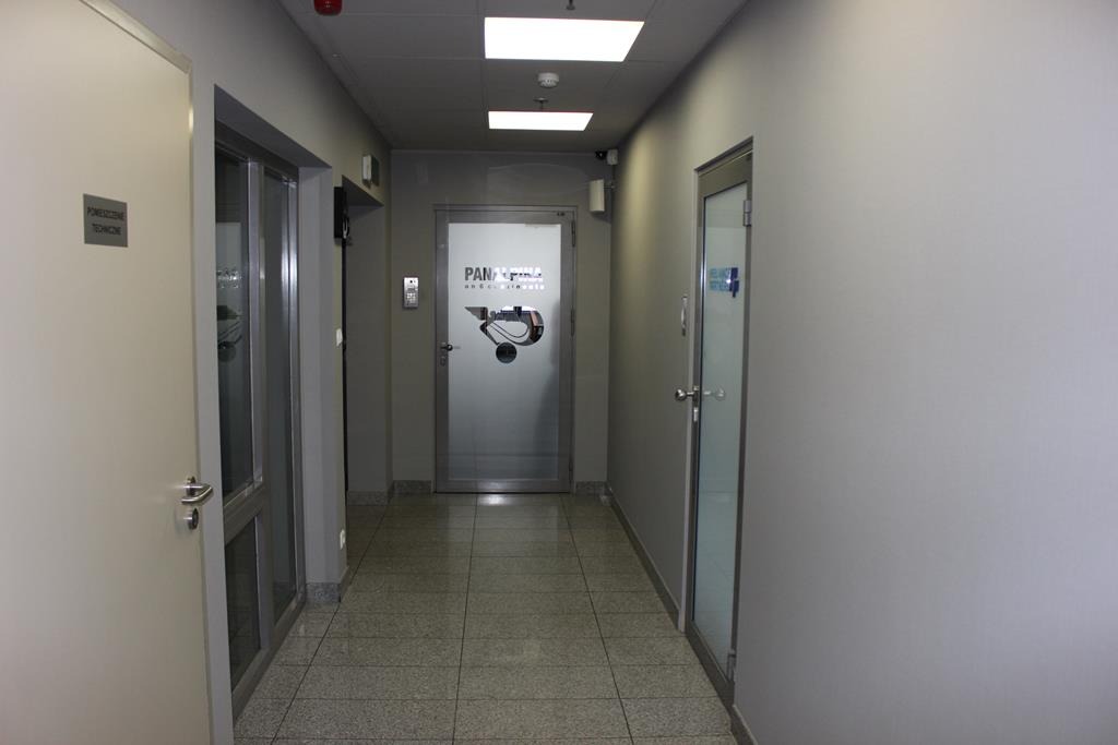 Korytarz biurowy z szarymi ścianami i drzwiami, widoczne logo firmy na szklanych drzwiach na końcu korytarza.