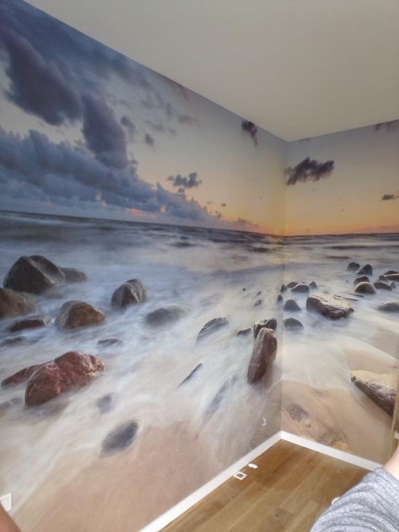 Fototapeta z motywem morza i kamieni na ścianie w Gdyni, imitująca plażę w pomieszczeniu z drewnianą podłogą.