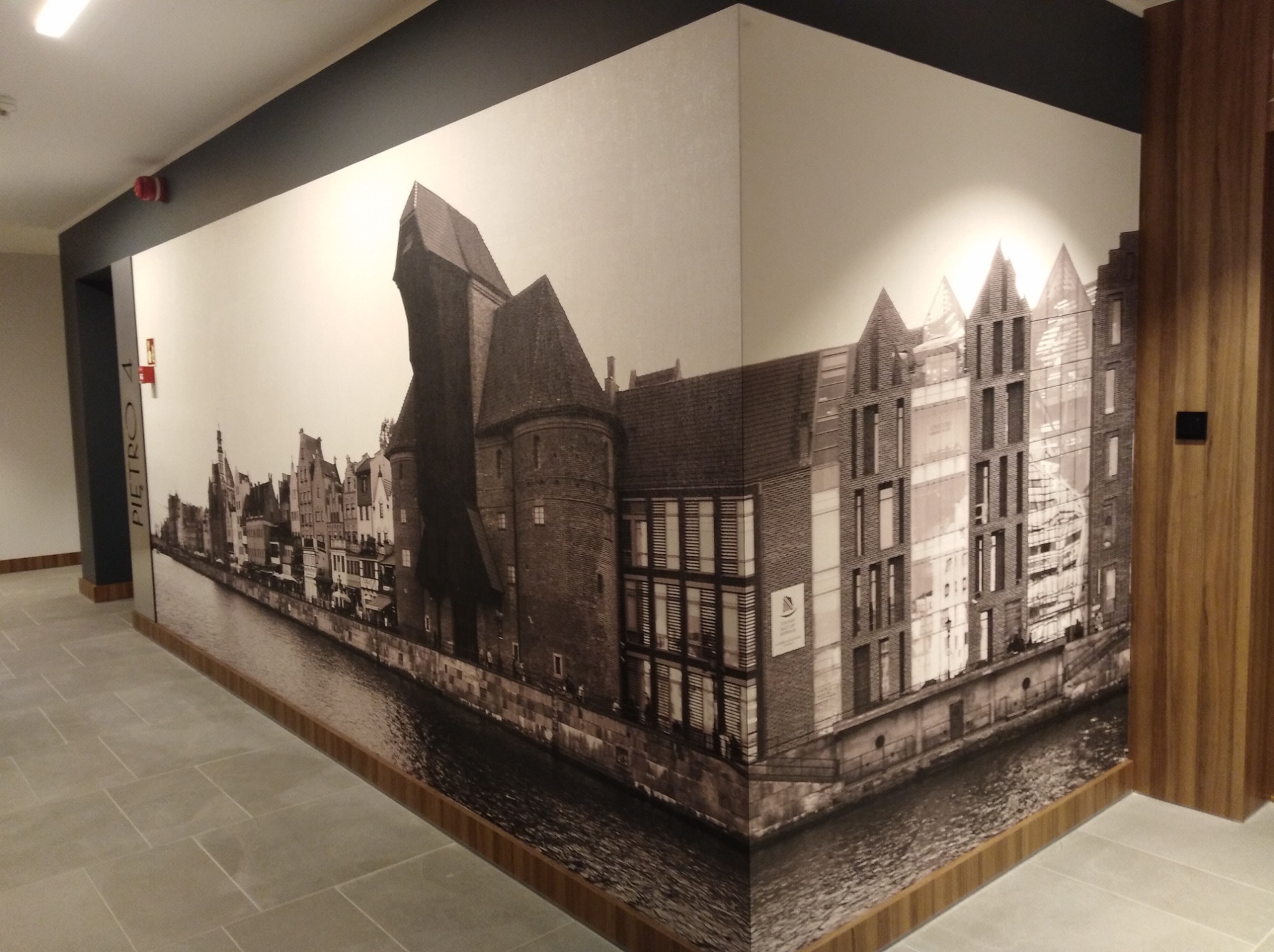 Fototapeta przedstawiająca panoramę Gdańska w czerni i bieli, umieszczona w korytarzu z szarą podłogą i drewnianymi wykończeniami.