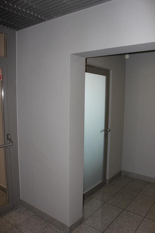 Świeżo odmalowana szara ściana z widocznym wejściem do pomieszczenia z matową szybą w stalowej ramie, płytki na podłodze z szarego lastryko.