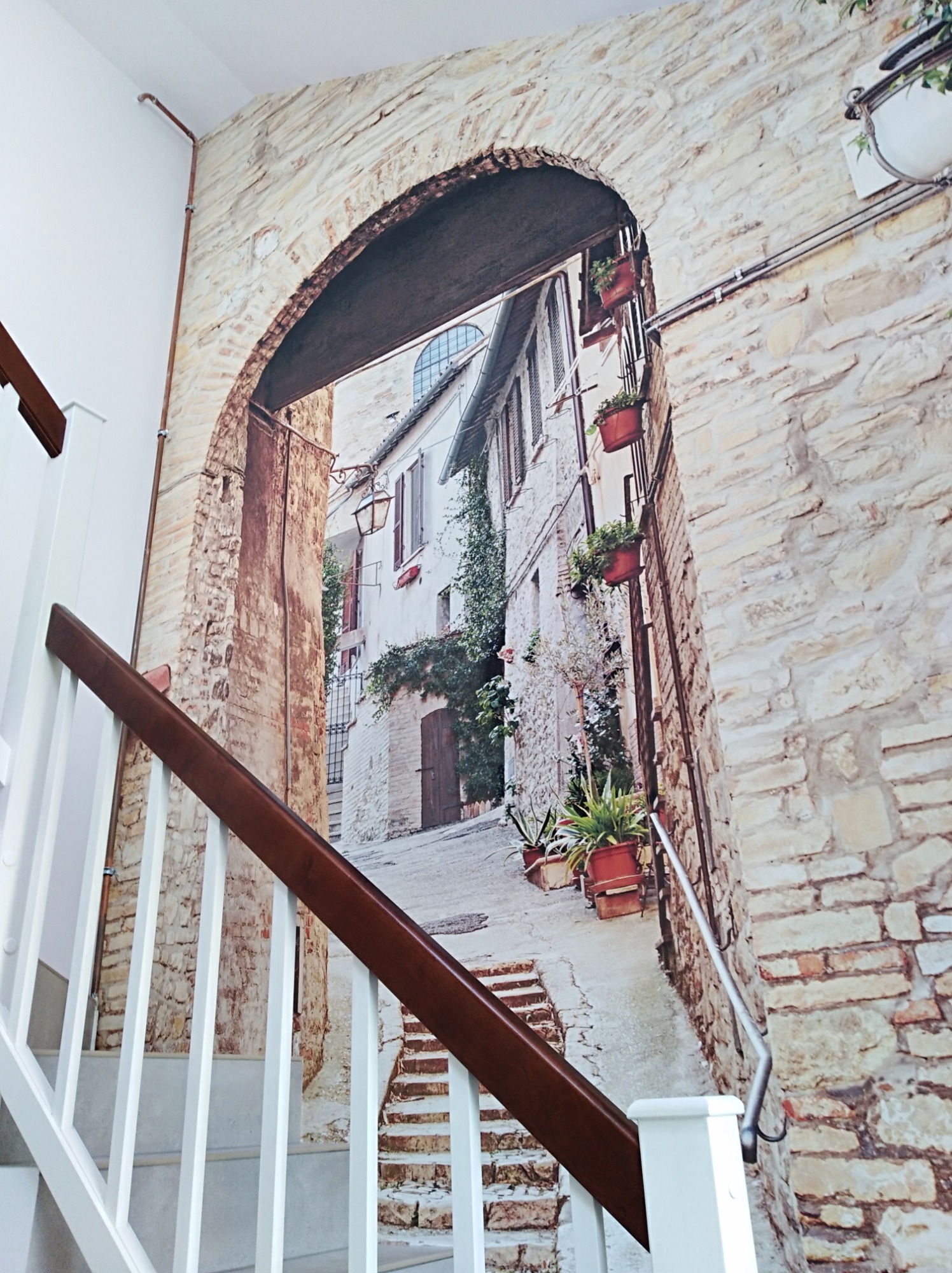Fototapeta z motywem toskańskiej uliczki z kamiennymi budynkami i doniczkami z kwiatami, zamontowana na ścianie klatki schodowej, widoczna fragment poręczy schodów.