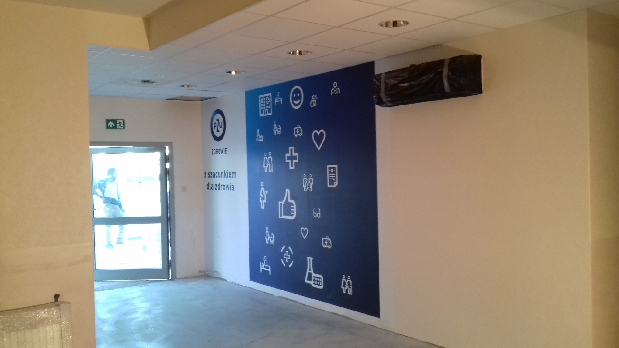 Świeżo wykończone wnętrze z dużą, niebieską grafiką na ścianie przedstawiającą białe ikony związane ze zdrowiem i logo PZU Zdrowie, widoczne drzwi ewakuacyjne i klimatyzator owinięty folią.