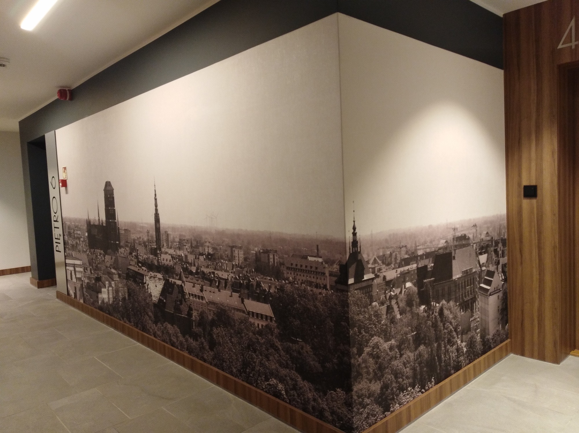 Korytarz z fototapetą przedstawiającą panoramę starego miasta w odcieniach szarości, z widocznymi wieżami kościołów i gęstą zielenią drzew. Fototapeta wykończona drewnianą listwą przypodłogową...