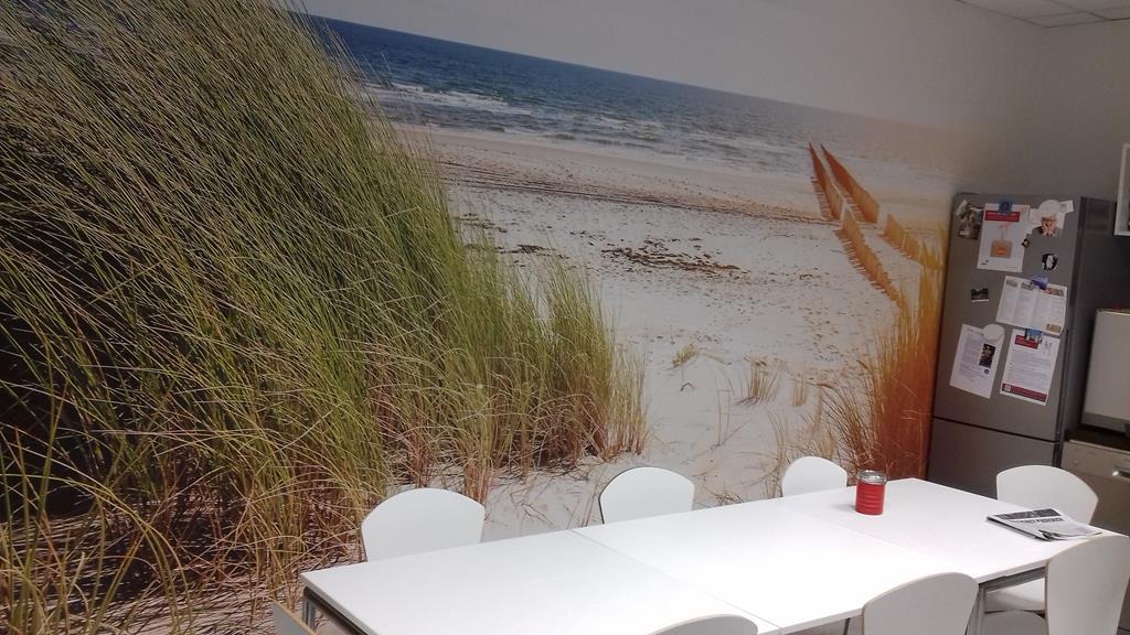 Fototapeta z motywem plaży z trawami morskimi na ścianie w biurze, widoczny stół konferencyjny z białymi krzesłami i lodówka z magnesami.