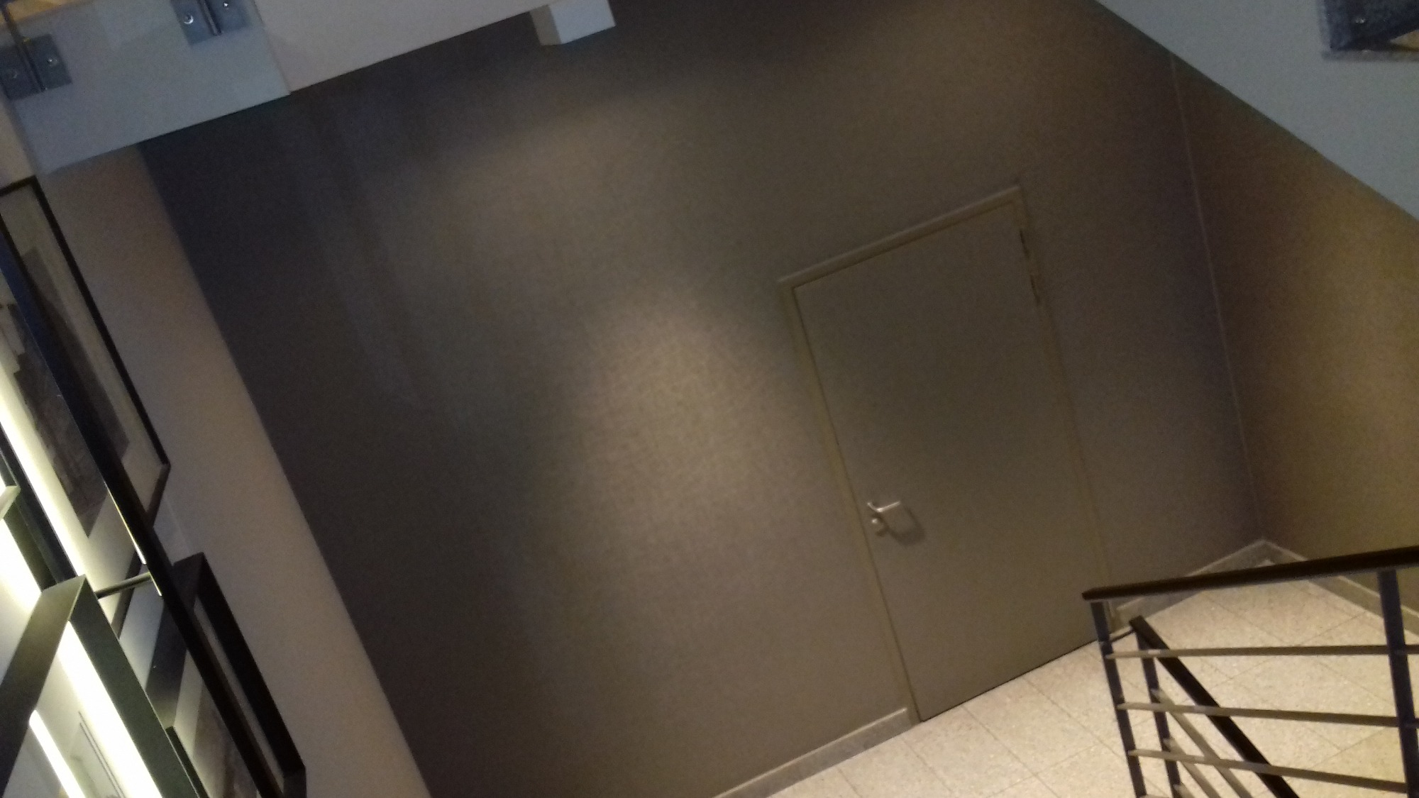 Ściana klatki schodowej pokryta tapetą w odcieniach szarości z delikatnym wzorem, obok drzwi i fragmentu balustrady.