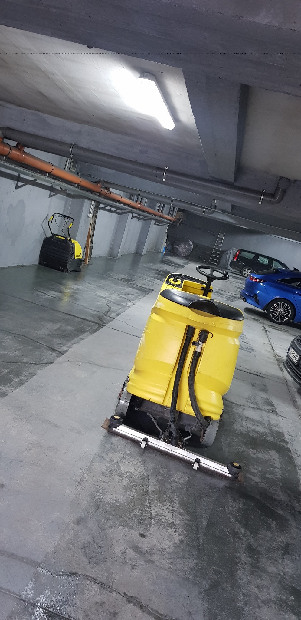 Żółta maszyna do mycia posadzek w garażu podziemnym, widoczny fragment umytej podłogi, w tle zaparkowane samochody.