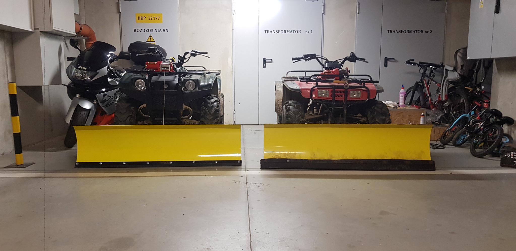 Dwa quady z zamontowanymi żółtymi pługami do odśnieżania stoją w garażu, obok motocykla i rowerów, na tle drzwi transformatorów.