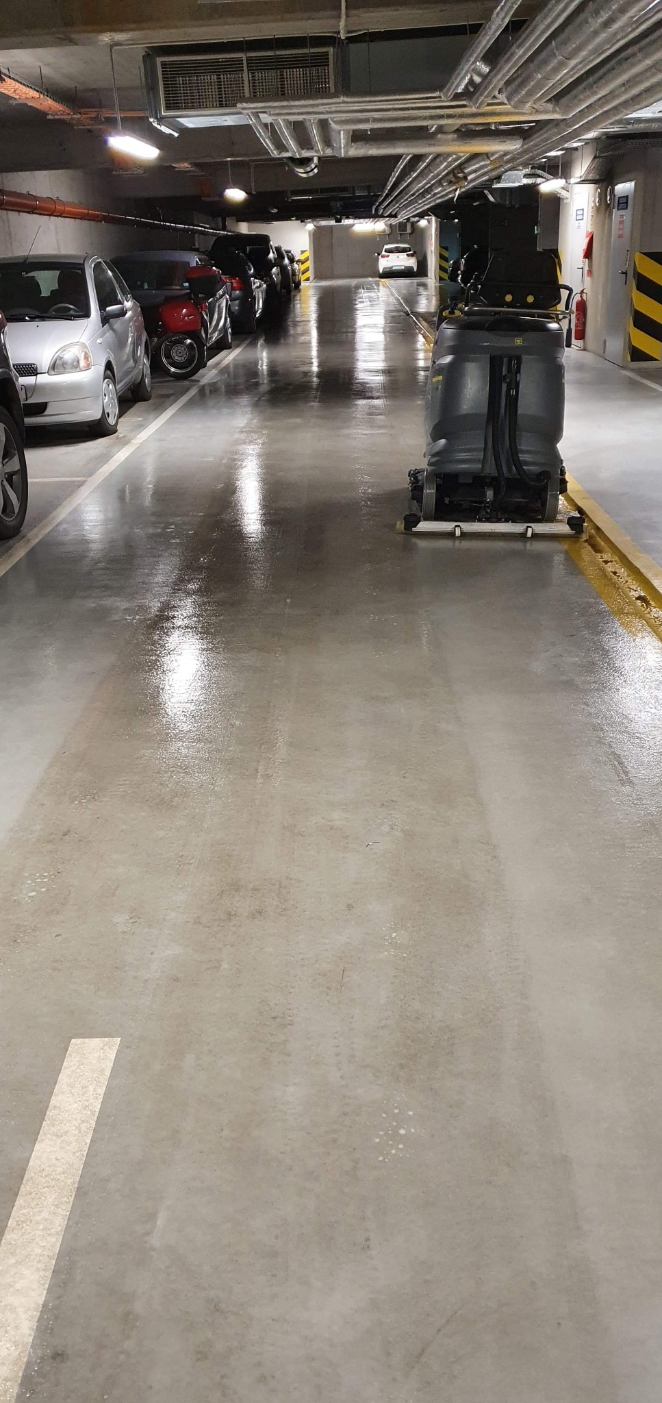 Automat do mycia podłóg w garażu podziemnym, mokra posadzka z widocznymi śladami czyszczenia, zaparkowane samochody w tle.