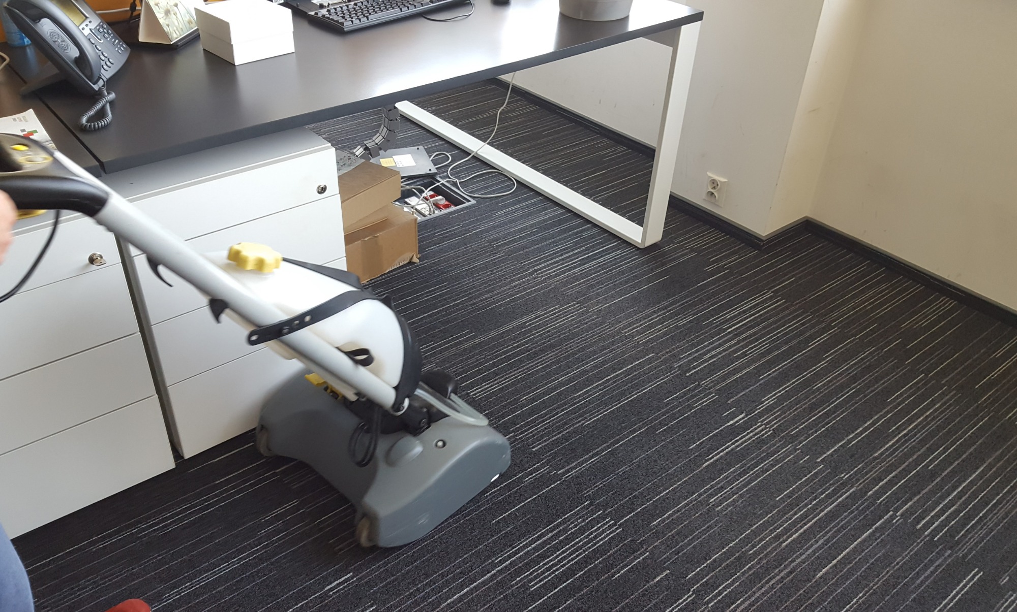 Maszyna do czyszczenia dywanów w biurze podczas pracy, widoczny fragment biurka i szafki, na podłodze dywan w ciemnych kolorach z jasnymi paskami.
