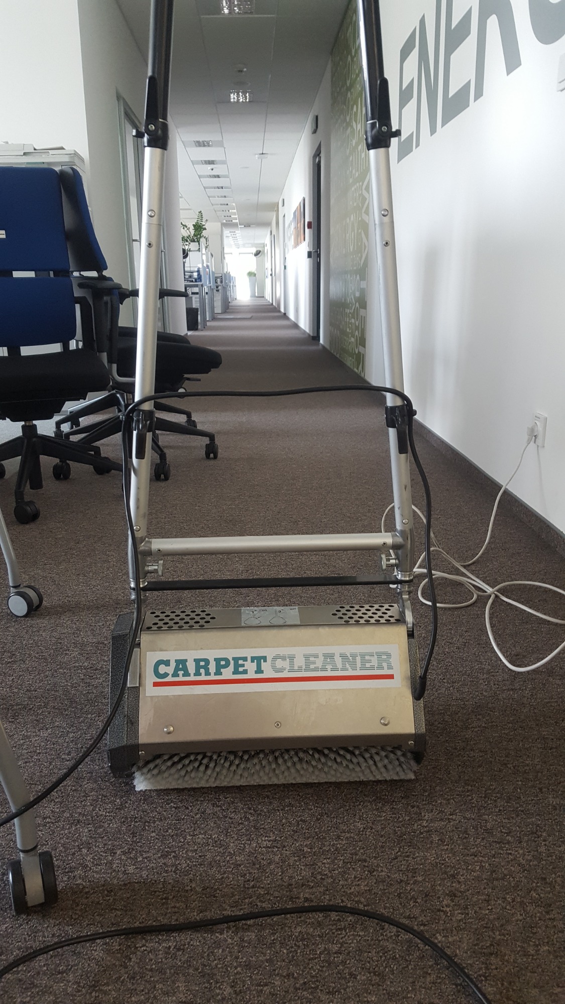 Profesjonalna maszyna do czyszczenia dywanów z napisem 'CARPET CLEANER' w biurowym korytarzu, widok z perspektywy operatora, z widocznymi krzesłami biurowymi i długim korytarzem w tle.