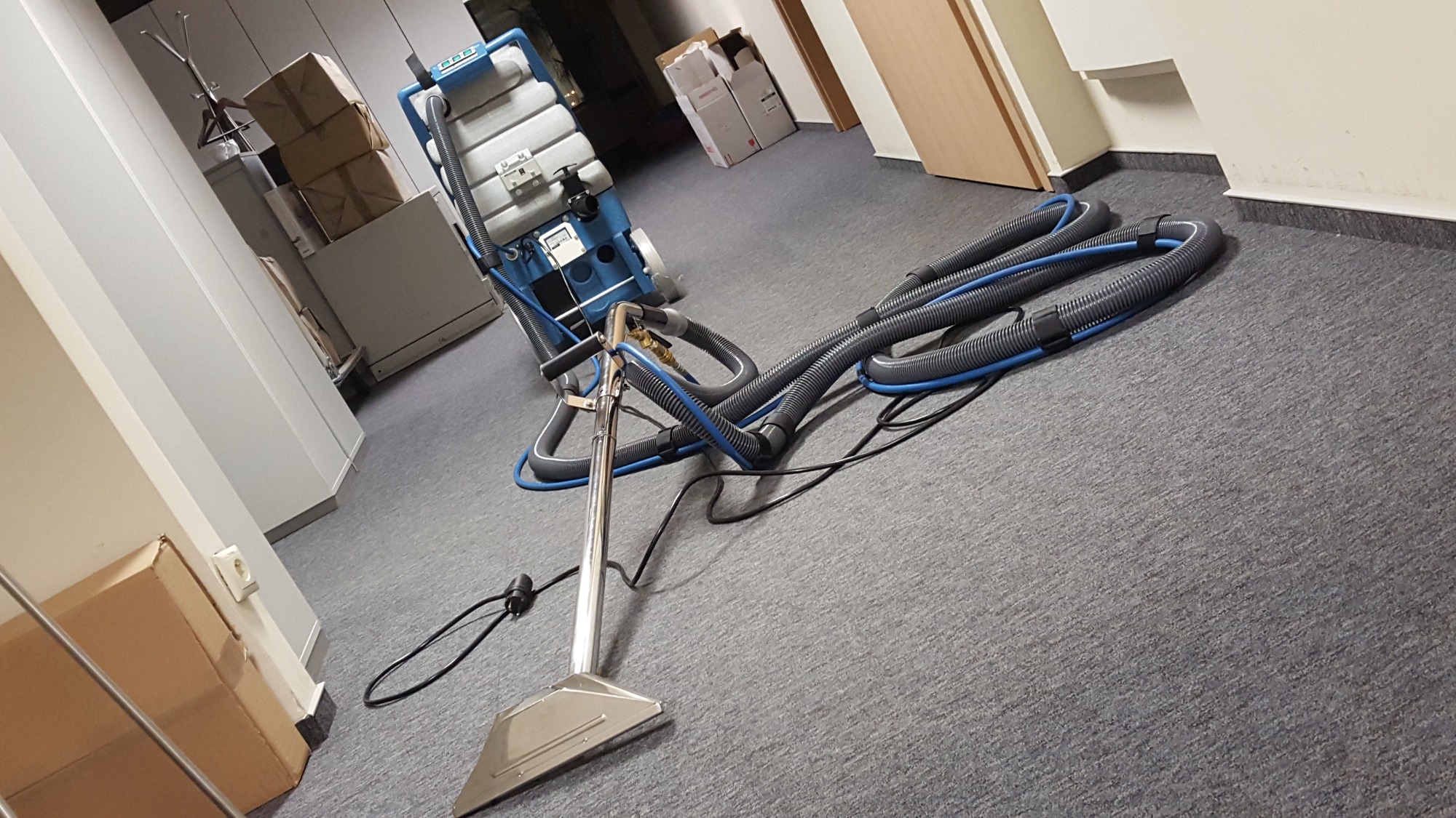 Profesjonalny odkurzacz piorący z długimi wężami na szarym dywanie w biurze, w tle widoczne kartony i szafy.