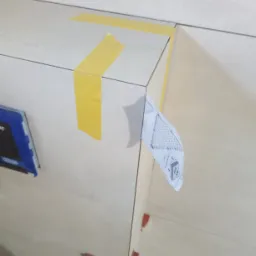 Fragment ściany z jasnych płytek ceramicznych w trakcie montażu, zabezpieczonych ż&oacute;łtą taśmą malarską na krawędziach, z użyciem plastikowych krzyżyk&oacute;w dystansowych i przyklejoną ulotką informacyjną...