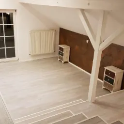 Poddasze po remoncie: jasne panele podłogowe z białymi listwami przypodłogowymi, ściana z efektem rdzy, biała stolarka okienna i drewniana konstrukcja nośna, dwie małe komody w stylu vintage.