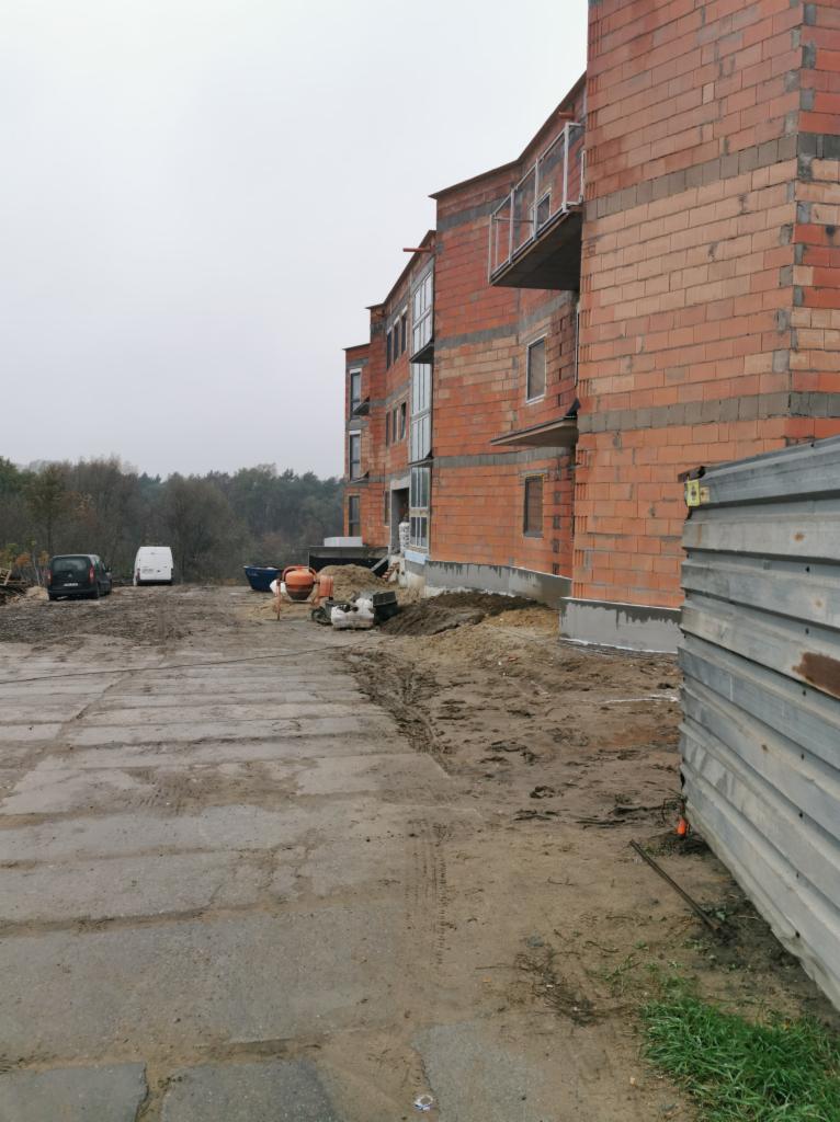 Wznoszenie wielorodzinnego budynku z czerwonej cegły, widoczne z zewnątrz, z częściowo zamontowanymi oknami i balkonami, na placu budowy z betonową drogą i stertami materiałów.