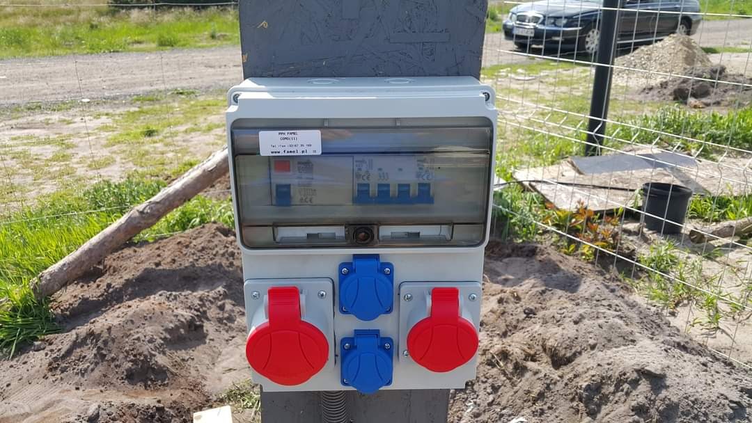 Zewnętrzna skrzynka elektryczna z bezpiecznikami i gniazdami siłowymi w obudowie z tworzywa sztucznego, zamontowana na słupku, widoczna tabliczka znamionowa i instalacja w ziemi.
