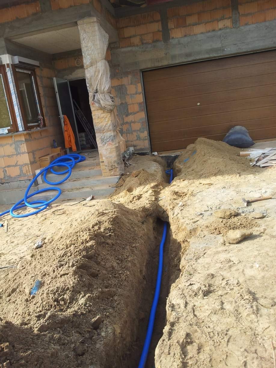 Niebieska rura w wykopie ziemnym przed budynkiem w trakcie budowy, widoczne cegły i brama garażowa.