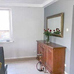 StoWnetrz - Szare ściany w pokoju po remoncie, drewniany kredens z bukietem czerwonych róż, widok na łazienkę w tle.