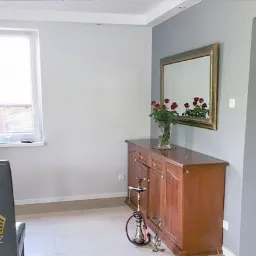 Szare ściany w pokoju po remoncie, drewniany kredens z bukietem czerwonych róż, widok na łazienkę w tle.