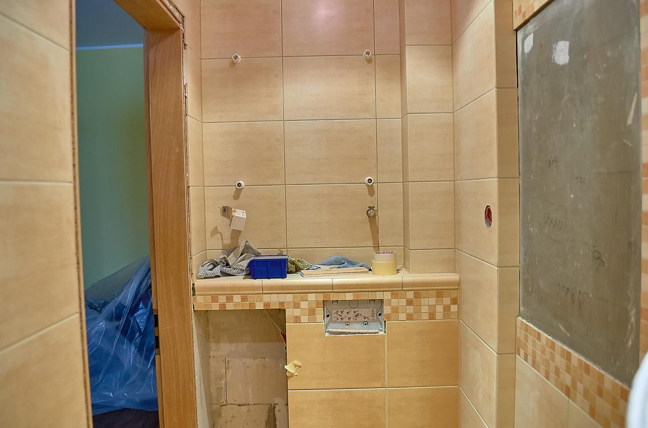 Łazienka w trakcie remontu: beżowe płytki na ścianach, częściowo wykończona zabudowa pod umywalkę z dekoracyjnym pasem z mozaiki, widoczne instalacje i narzędzia, ściana z zaznaczonymi wymiarami.