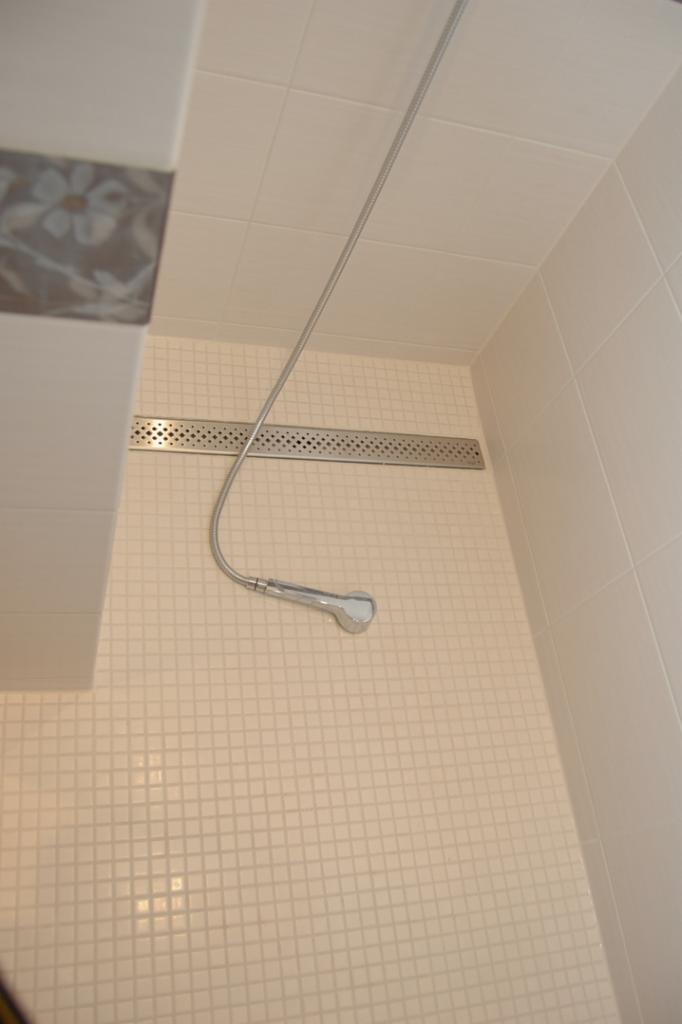 Wykończona kabina prysznicowa z mozaiką w odcieniach bieli i beżu, z odpływem liniowym i słuchawką prysznicową na elastycznym wężu.