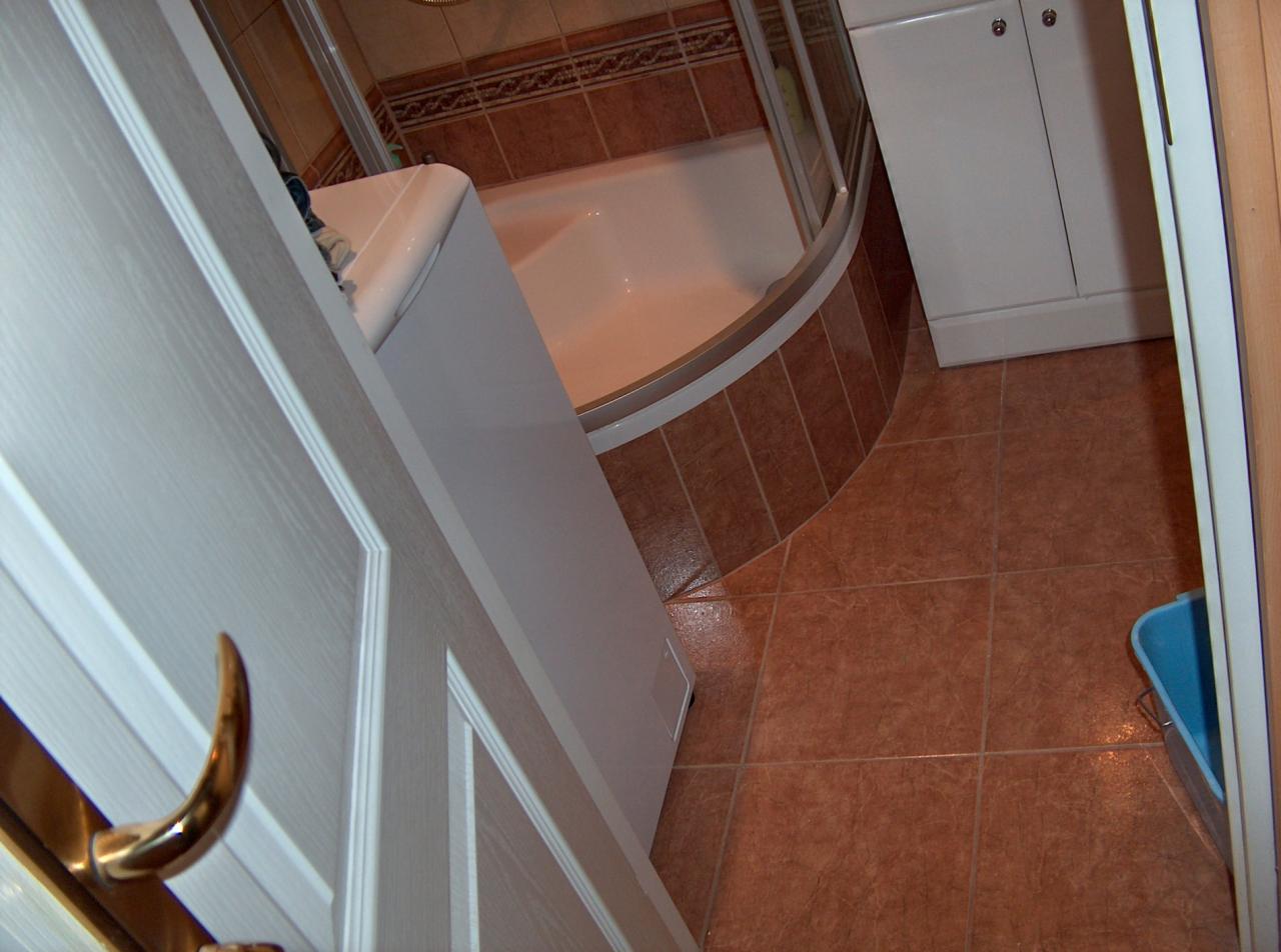 Widok z góry na wykończoną łazienkę z brązowymi płytkami, białą wanną z kabiną prysznicową i szafką, widzianą przez uchylone białe drzwi z pozłacaną klamką.