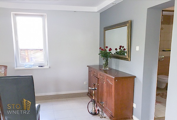 Szare ściany w pokoju po remoncie, drewniany kredens z bukietem czerwonych róż, widok na łazienkę w tle.