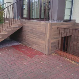 Wykończenie elewacji budynku z brązowymi płytkami imitującymi drewno, schody wejściowe z metalową balustradą i czerwona kostka brukowa na podwórku.