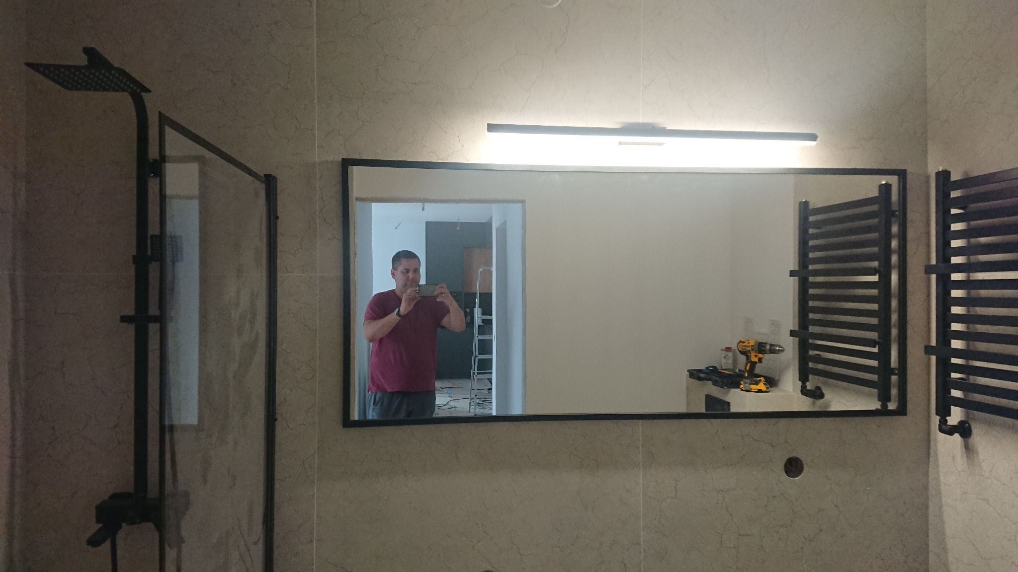 Nowoczesna łazienka z czarną armaturą: kabina prysznicowa typu walk-in, grzejnik drabinkowy i lustro z oświetleniem. W lustrze odbija się osoba robiąca zdjęcie oraz drabina. Na blacie w odbiciu...