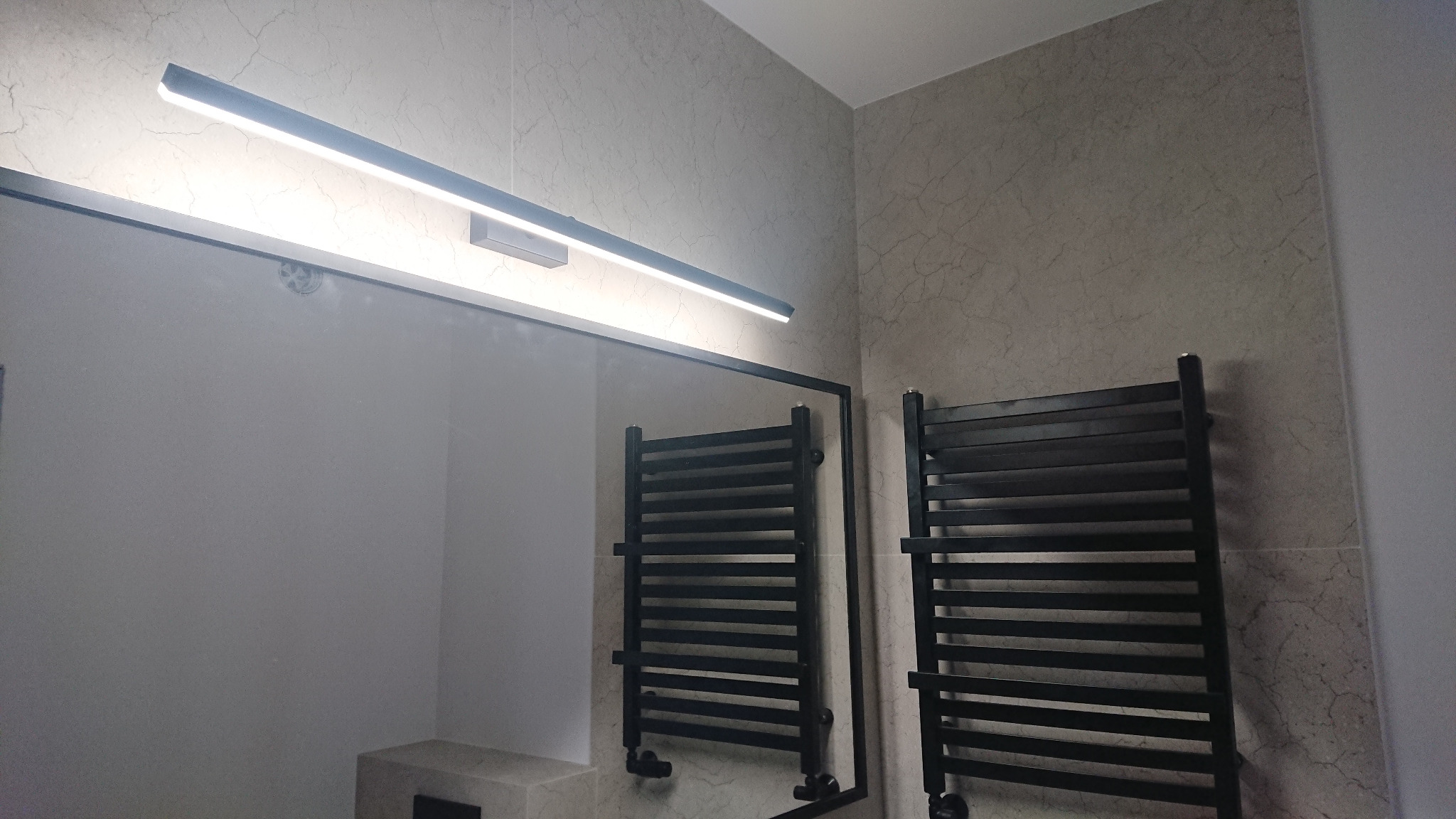 Nowoczesna łazienka z grzejnikiem drabinkowym w czarnym kolorze, podświetlanym lustrem i płytkami imitującymi marmur.