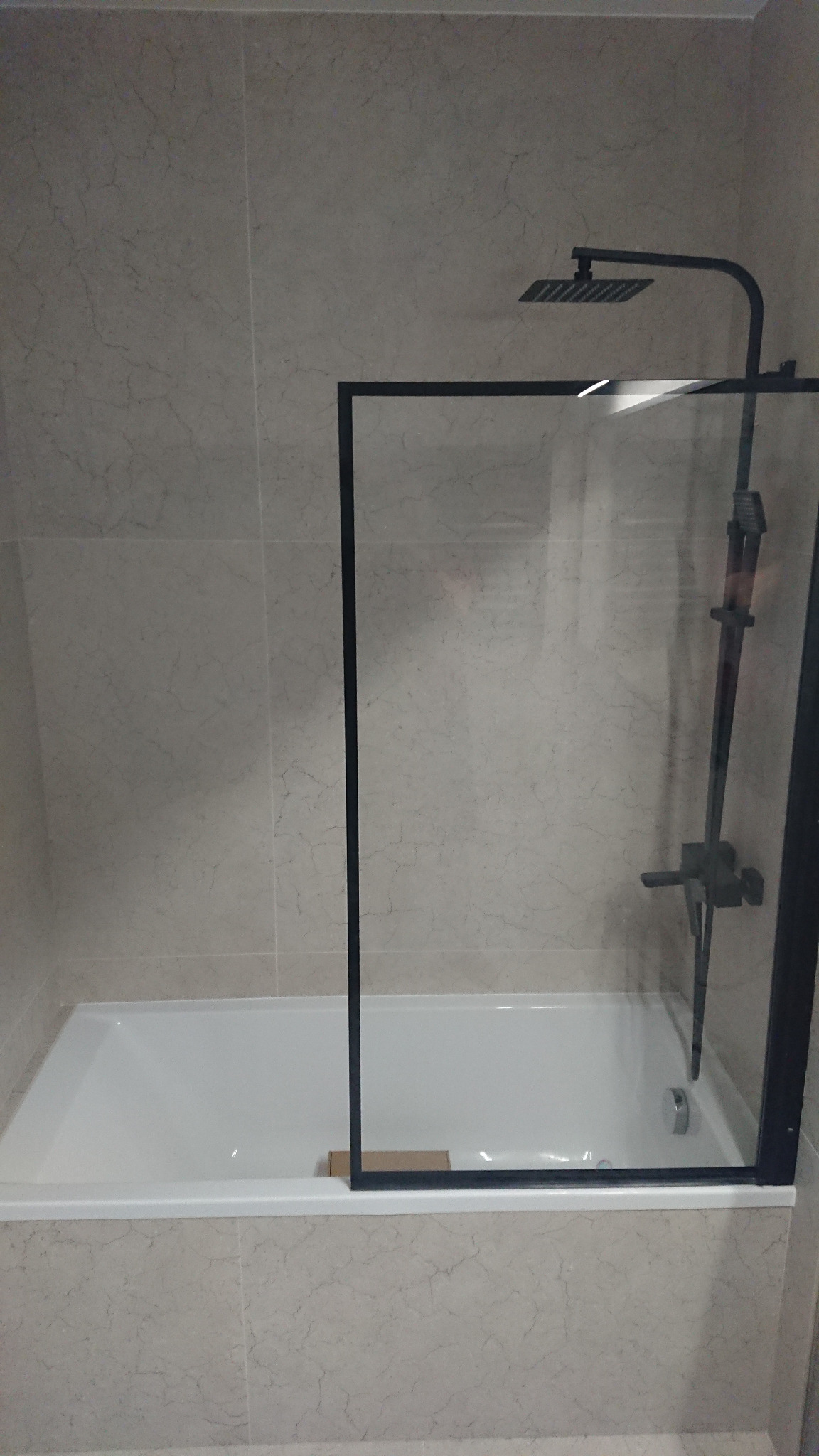 Biała wanna z czarną szklaną ścianką prysznicową i czarną armaturą na tle beżowych płytek imitujących marmur.