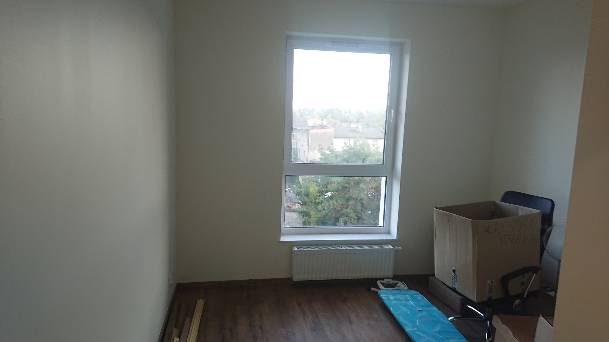 Pusty pokój po remoncie z drewnopodobną podłogą, jasnymi ścianami, białym oknem z widokiem na budynki i zieleń, grzejnikiem pod oknem oraz kartonem z napisem i krzesłem w rogu.
